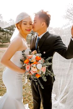 Side view of Asian groom kissing bride with bouquet during romantic wedding celebration standing under veil