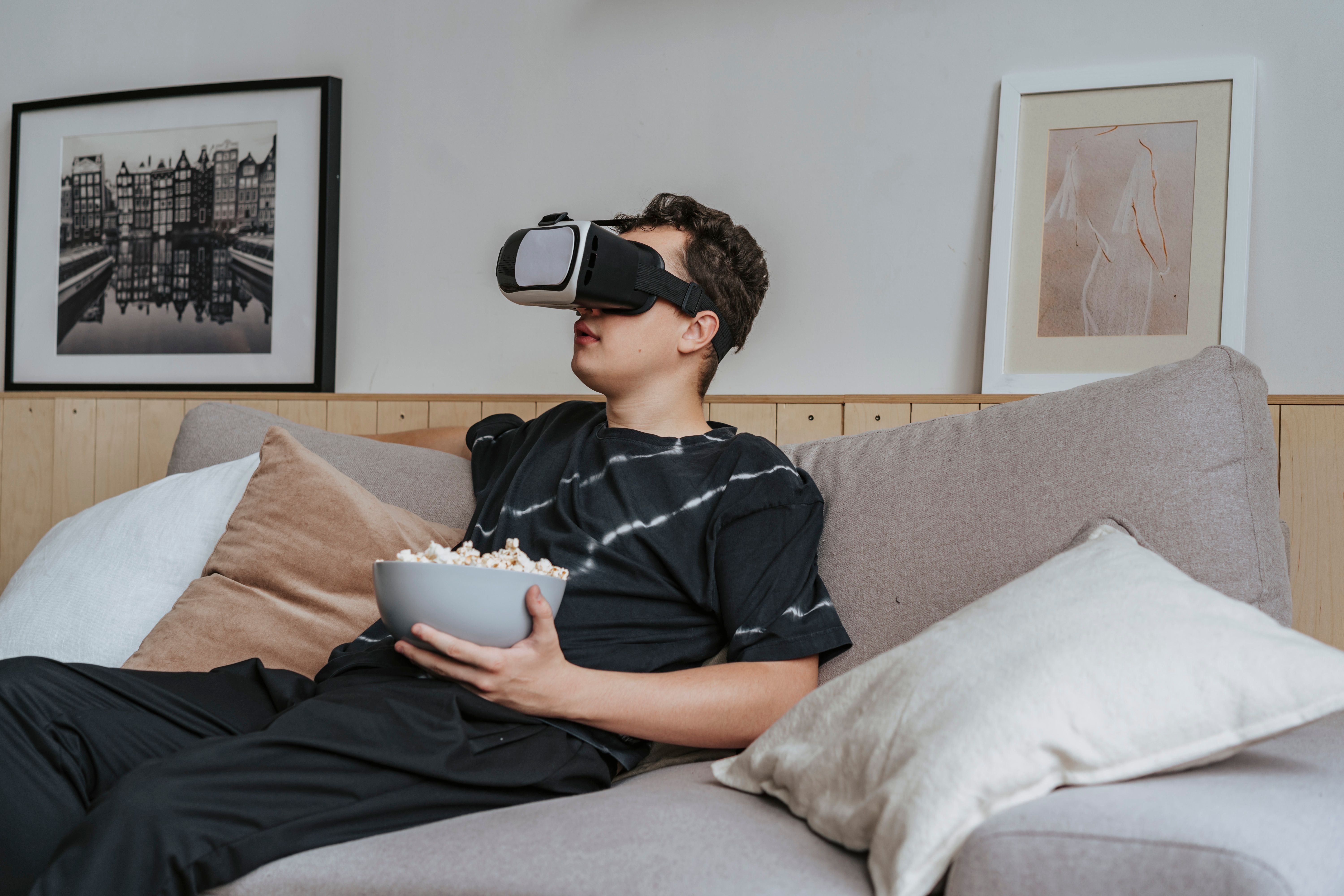 A young man in a black shirt enjoys virtual reality while seated on a sofa with a bowl of popcorn.