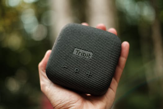 Hand holding a Tribit portable speaker with blurred outdoor background, featuring modern design and textured surface.