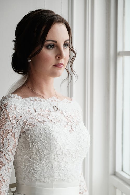 Elegant bride with trendy hairstyle wearing classy white dress standing in light room and looking out window