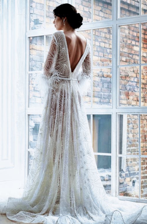 Full length back view gorgeous bride wearing luxurious translucent white dress with train standing near big window in light modern studio