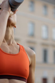 Fit woman in an orange sports bra drinking from a water bottle outdoors, symbolizing fitness and healthy lifestyle.