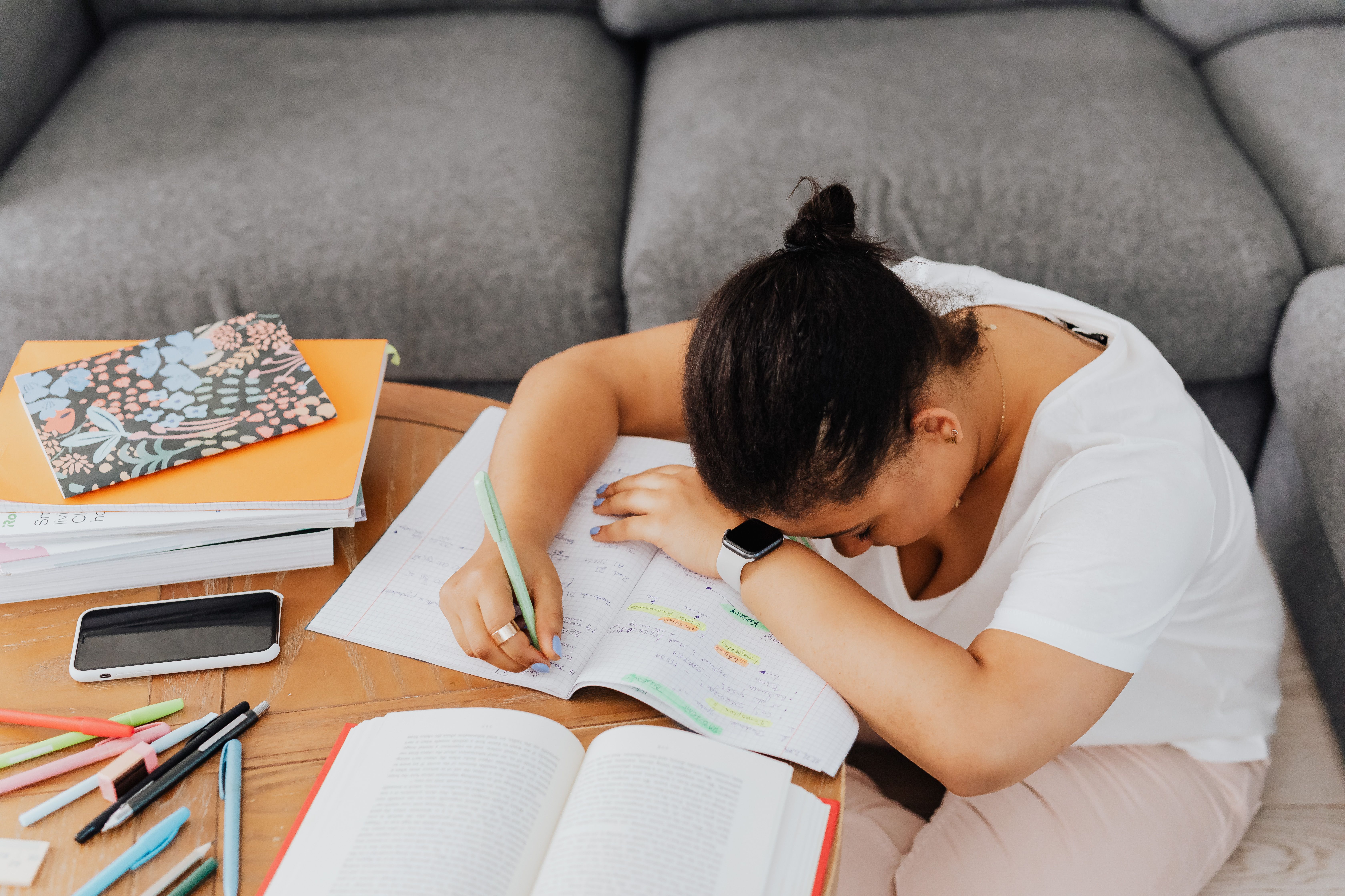 Young woman in a white shirt looking tired and resting on a couch while surrounded by study materials, notebooks, and pens.