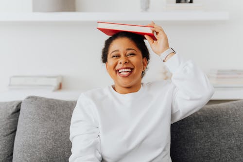 A Laughing Woman in White Sweater Holding a Book on Top of Her Head