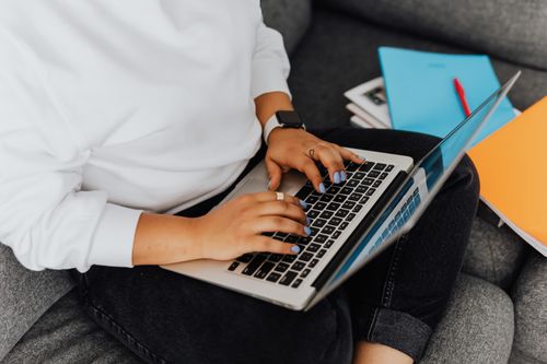 Free Person using a laptop on a gray sofa, surrounded by colorful notebooks, concept of remote work or study. Stock Photo