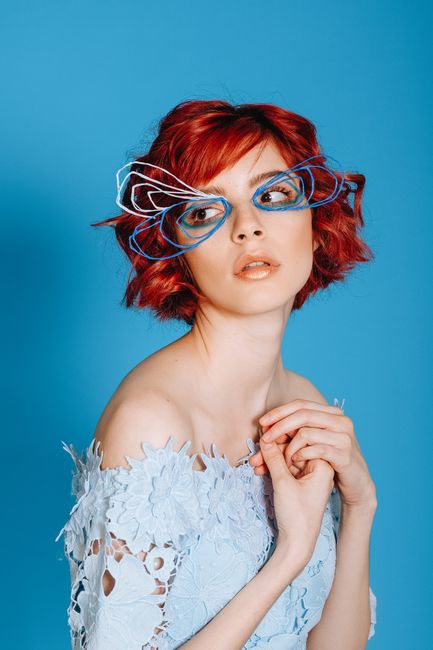 Gentle young female wearing lace dress with bare shoulders and creative wire face decoration while standing on blue background and looking away