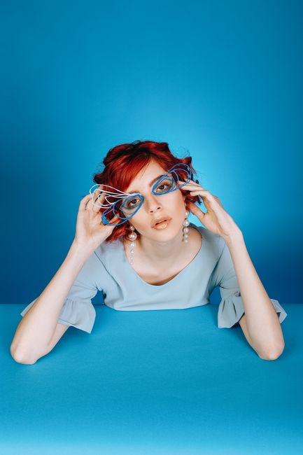 Young calm female in blue dress and creative wire glasses touching face tenderly while leaning on blue surface in studio and looking at camera