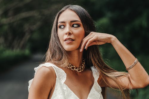 Charming female in white outfit and massive necklace touching cheek and looking away while spending warm summer day in green garden