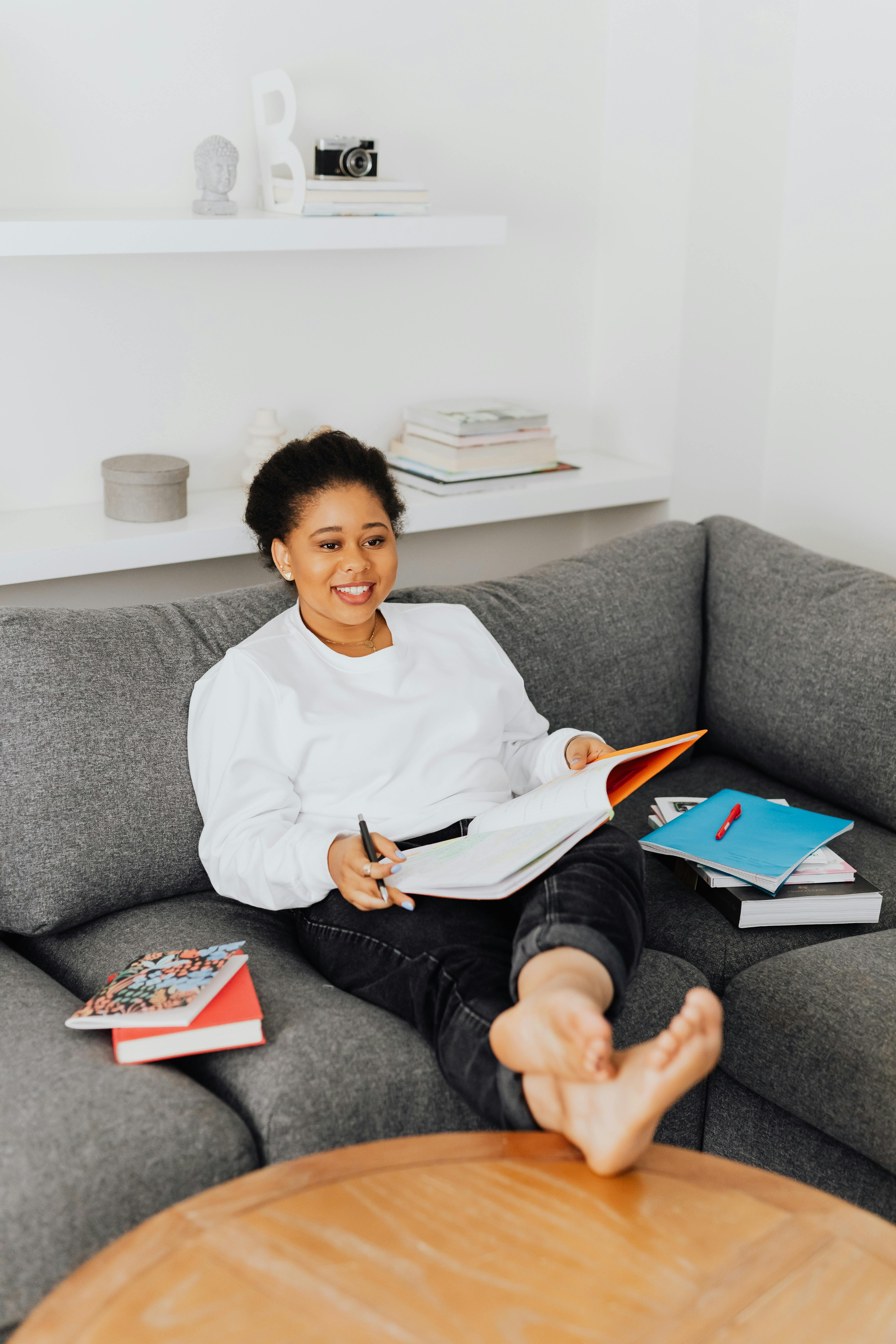 Black woman enjoying a study session at home, surrounded by books on a comfortable grey sofa.