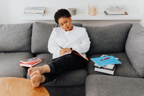 Free A young woman relaxes on a sofa, writing in a notebook surrounded by books. Stock Photo