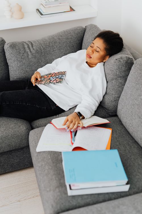 Free An exhausted student rests on a couch surrounded by books and notes. Stock Photo
