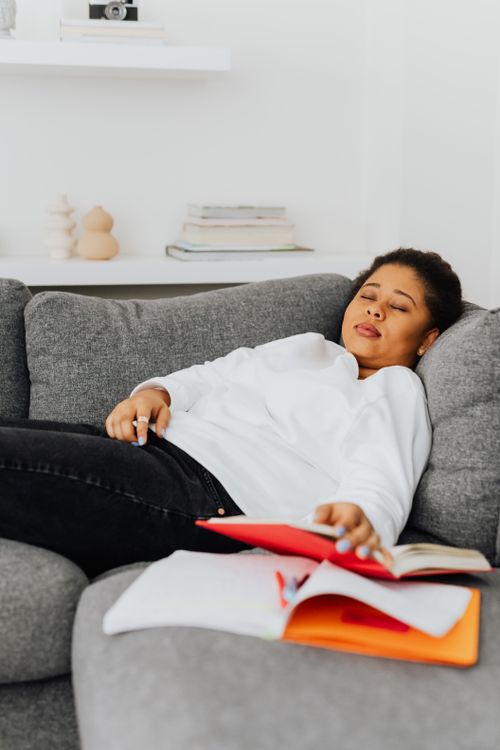Free An adult woman resting on a sofa surrounded by open books, reflecting study fatigue. Stock Photo