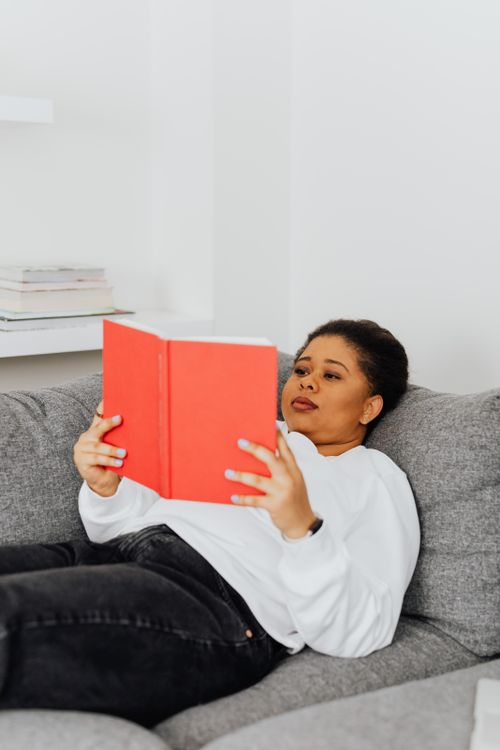 Free African American woman relaxing indoors, reading a book on a grey sofa. Stock Photo