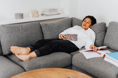 Free A young woman resting on a cozy grey sofa with open books around her, appearing tired after studying. Stock Photo