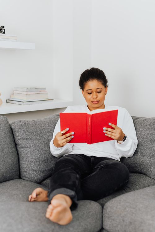 Free Black woman reading on a sofa at home, enjoying a relaxed afternoon. Stock Photo