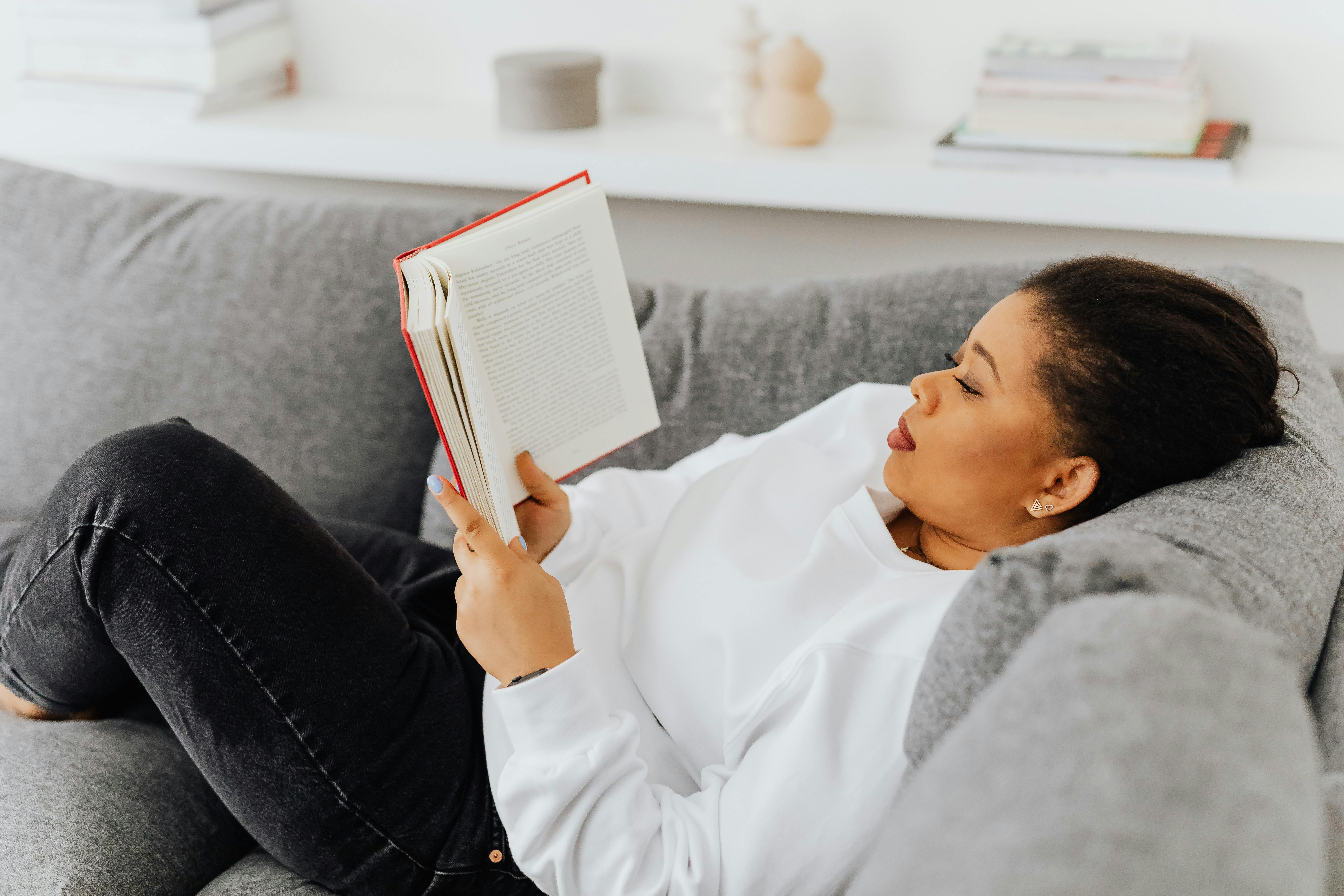 Free An adult woman enjoys a quiet reading session on a cozy gray couch indoors. Stock Photo