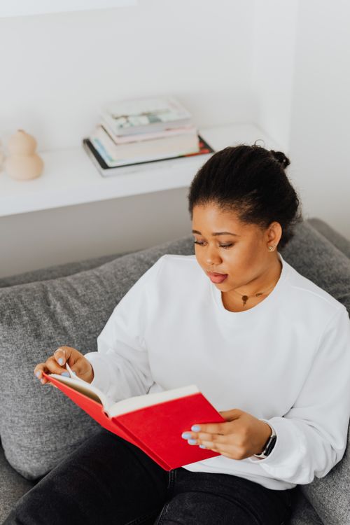 Free A woman in a white sweater reading a red book while sitting on a gray couch indoors. Stock Photo
