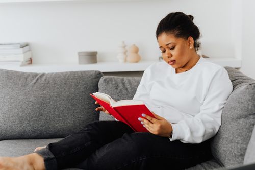 Free African American woman wearing casual attire reading a book on a comfortable sofa indoors. Stock Photo