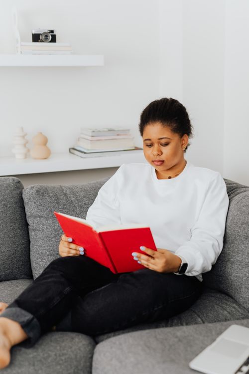 Free A woman in casual attire reading a red book on a cozy gray sofa indoors. Stock Photo