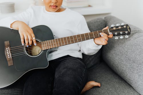 A Woman in White Long Sleeves Sitting on a Gray Couch while Strumming the Guitar