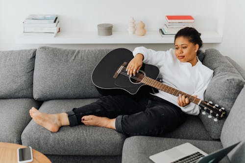 A Woman in White Sweater Sitting on the Couch while Playing Guitar
