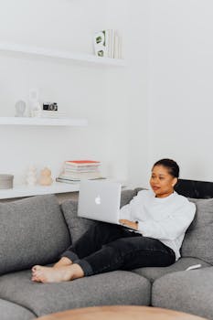 Woman using a laptop on a sofa in a stylish living room with shelves and decor.