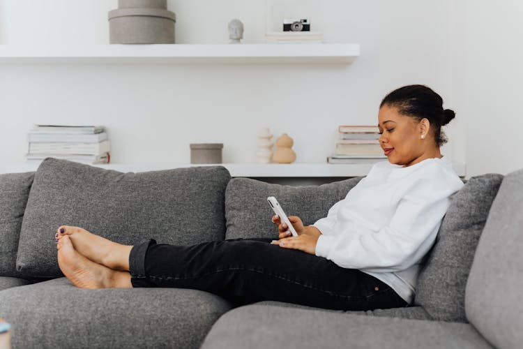 A Woman Sitting On The Couch While Using Her Mobile Phone