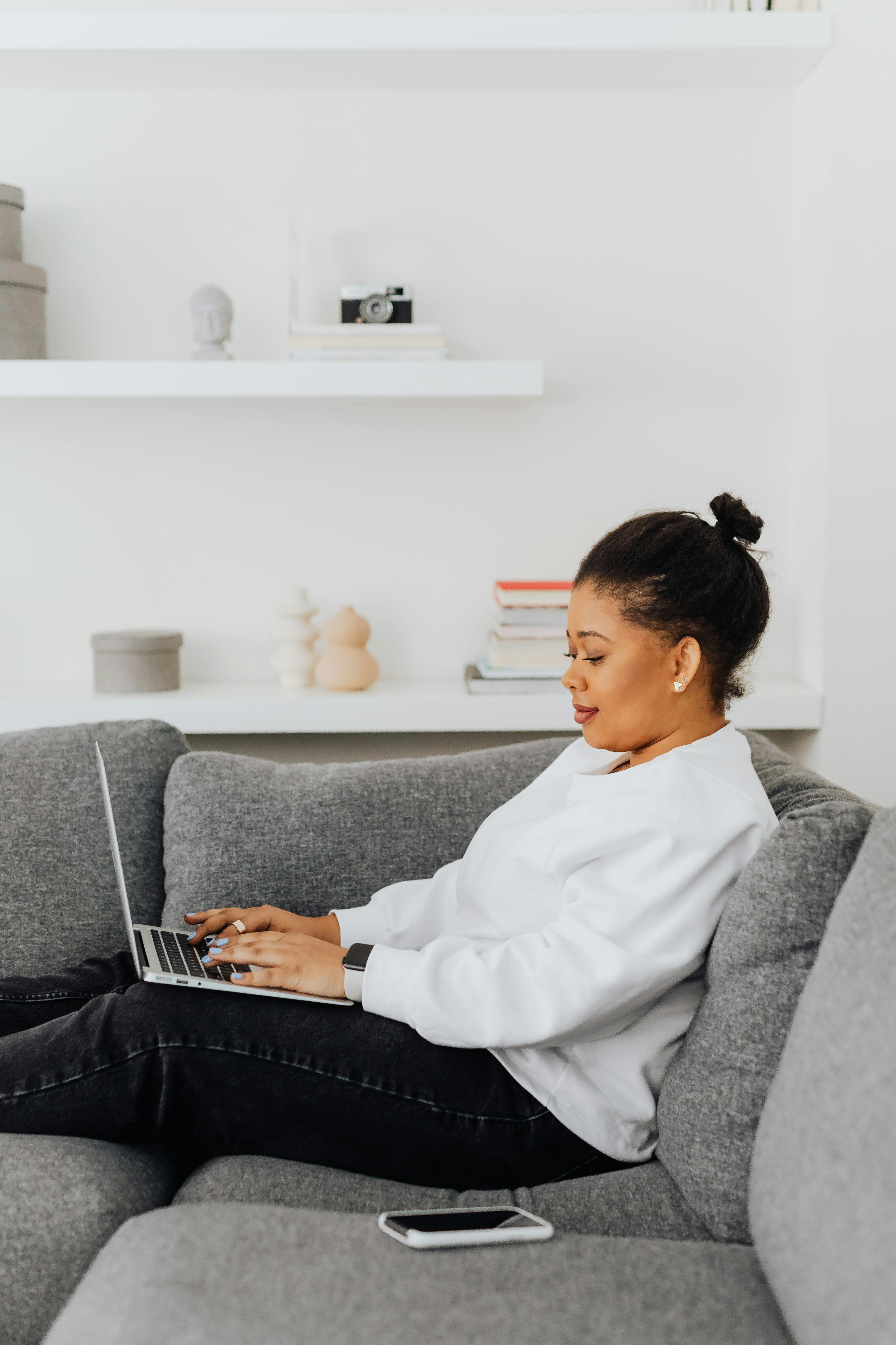 a woman in white sweater sitting on the couch while using her laptop