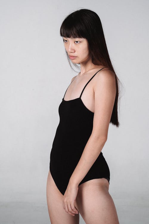 Free Unemotional young Asian female wearing black bodysuit standing confidently against white wall in studio and looking away Stock Photo