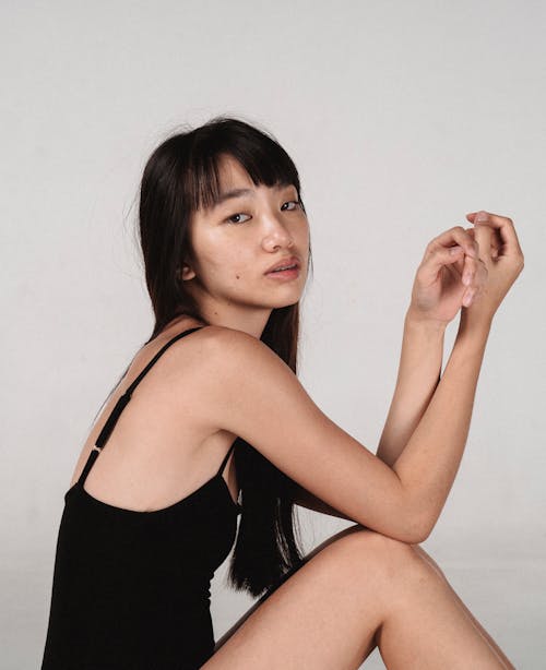 Side view young calm Asian female model wearing black bodysuit sitting against white background in studio and looking at camera