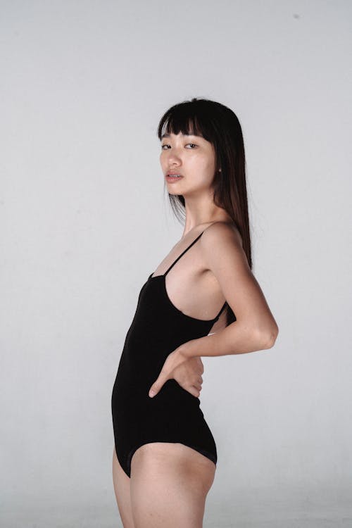Side view slender young Asian female model in tight black bodysuit standing with hands on waist against white wall in light studio and looking at camera