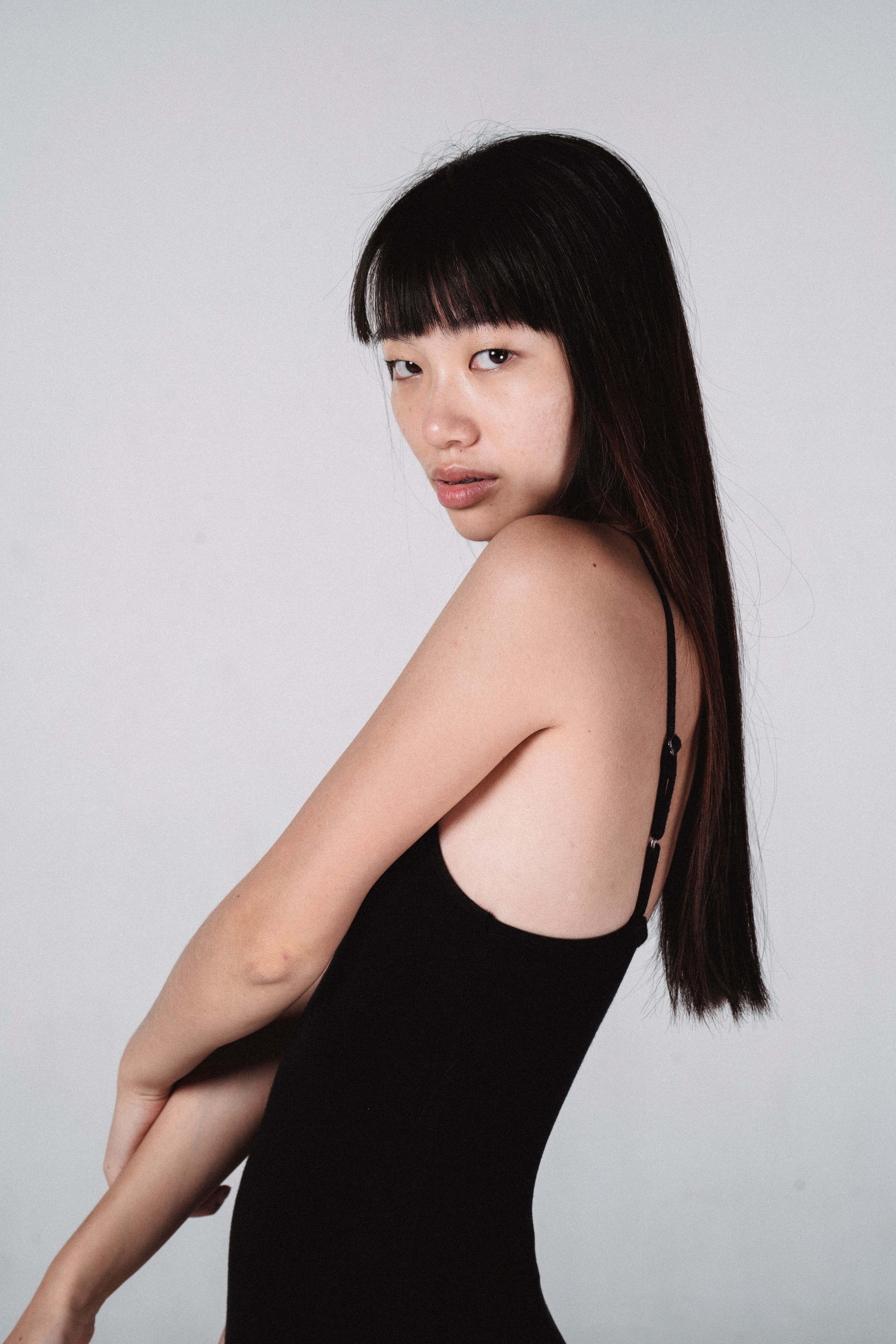 Side view appealing Asian female in tight black top standing against white wall and looking at camera sensually