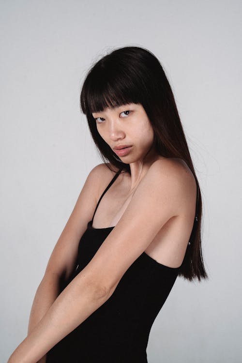 Free Side view sensitive young Asian female model wearing tight black top standing against white wall and looking at camera Stock Photo