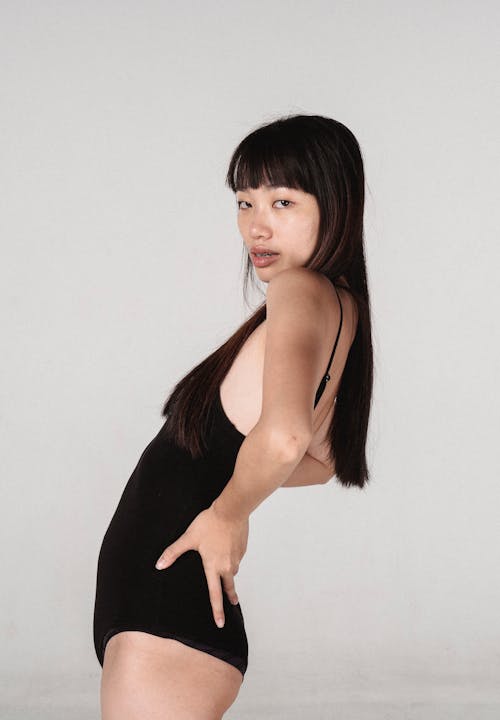 Side view attractive Asian female model in black bodysuit standing with hands on waist and bending back slightly while looking at camera in light studio