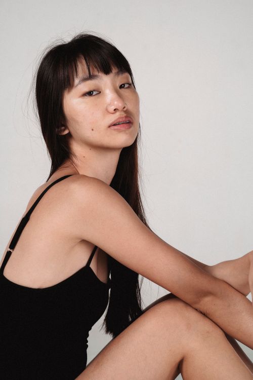 Free Side view unemotional charming Asian female model in tight black bodysuit sitting against light background in modern studio and looking at camera Stock Photo