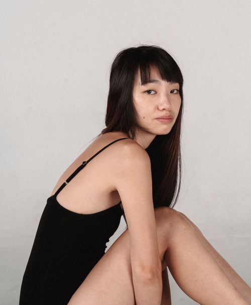 Free Side view of young pensive ethnic female model in black bodysuit sitting against gray background and looking at camera Stock Photo