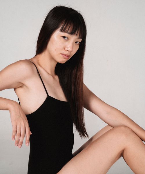 Free Side view of young focused ethnic woman in black bodysuit looking at camera while sitting near wall Stock Photo