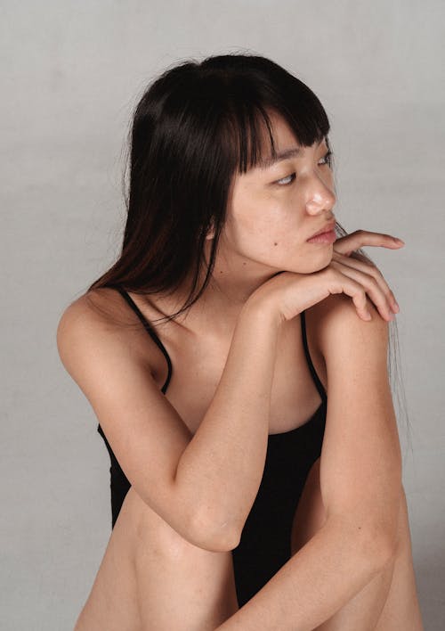 Young pondering Asian female model in underwear touching shoulder while leaning on hand and looking away on gray background