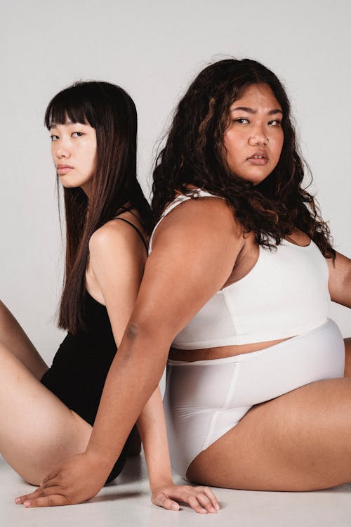 Free Side view of serious young plus size and slim ethnic females in seamless underwear sitting on floor back to back and looking at camera Stock Photo