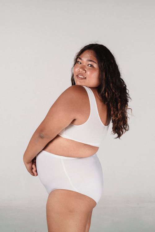 Free Side view of self assured young ethnic overweight female model in white underwear smiling and looking over shoulder while standing against gray background Stock Photo