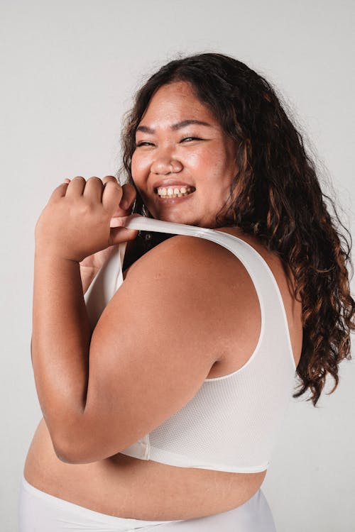 Side view of plus size Asian female pulling strap of bra and smiling on white background