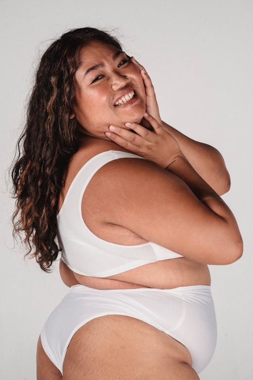 Side view of cheerful Asian woman with long curly hair in light underwear touching face and standing in studio