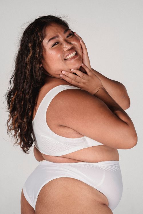 Free Side view of cheerful Asian woman with long curly hair in light underwear touching face and standing in studio Stock Photo