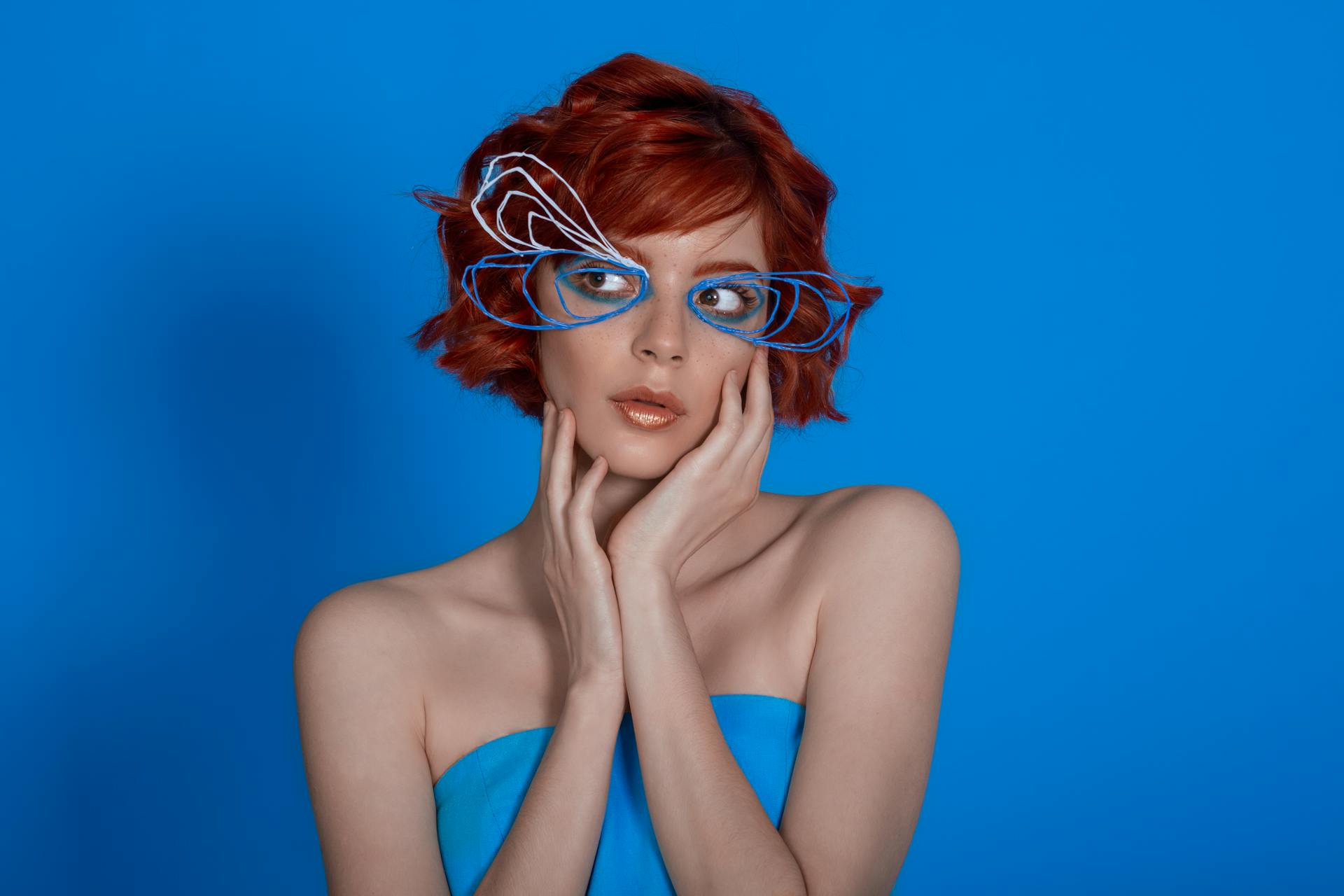 Creative studio portrait of a woman with red hair and unique eyewear against a blue backdrop.