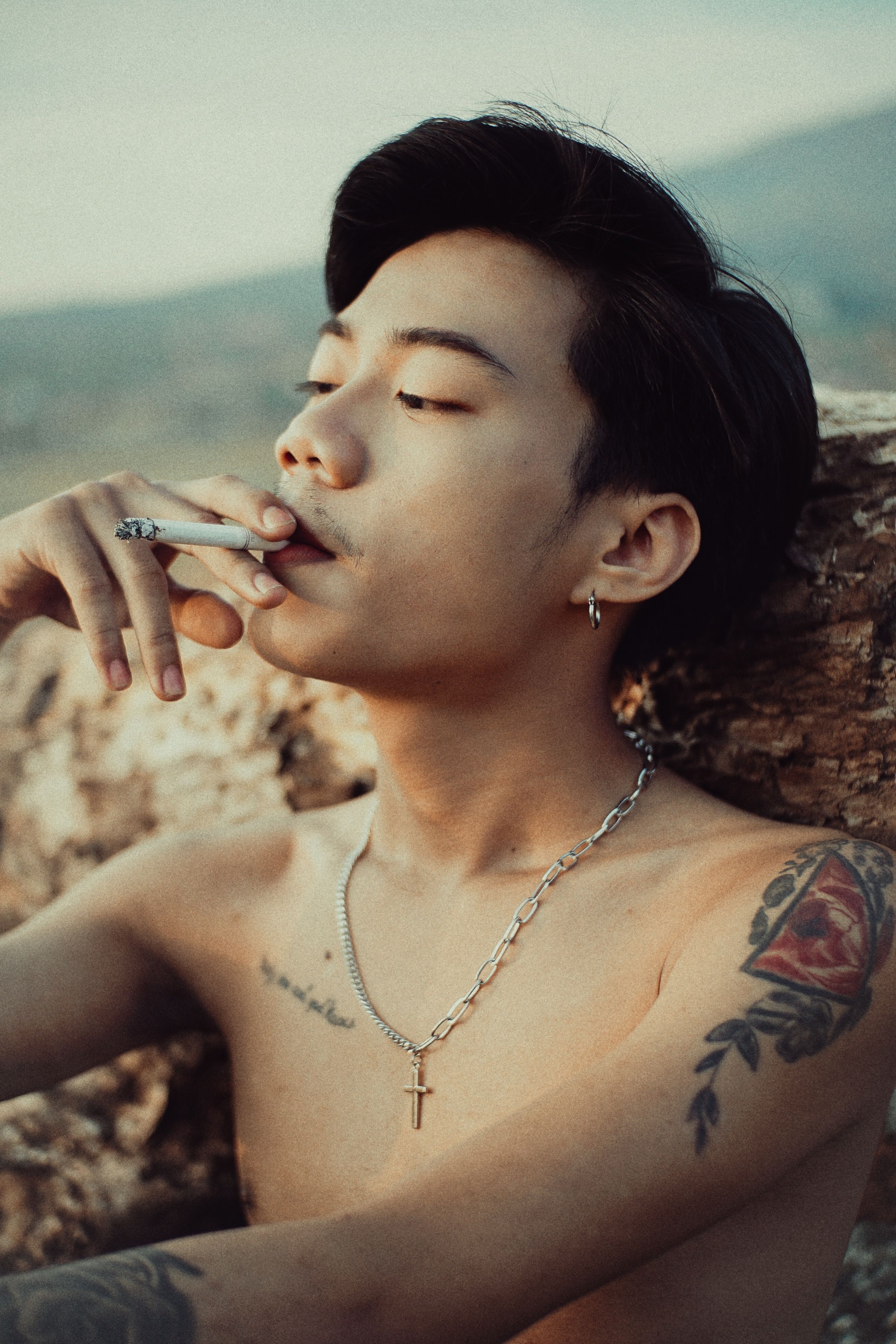 Portrait of a shirtless man smoking a cigarette outdoors, displaying tattoos.