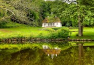 宁静的湖泊被郁郁葱葱的绿树环绕，湖面上倒映着一座风景如画的小屋，茅草屋顶清晰可见