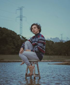 侧面图中，一位穿着休闲装、赤脚的中性人士，正坐在宁静乡村流淌的浅溪旁一张破旧的凳子上，沉思着望向镜头