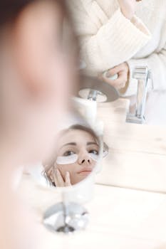 Close-up reflection of a woman applying under-eye patches in a round mirror, wearing a cozy white sweatshirt.