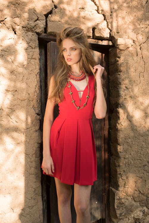 Woman in Red Sleeveless Dress Standing Beside a Wooden Door