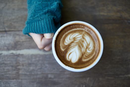 Cozy shot of a person holding a creatively styled cup of latte on a wooden table.